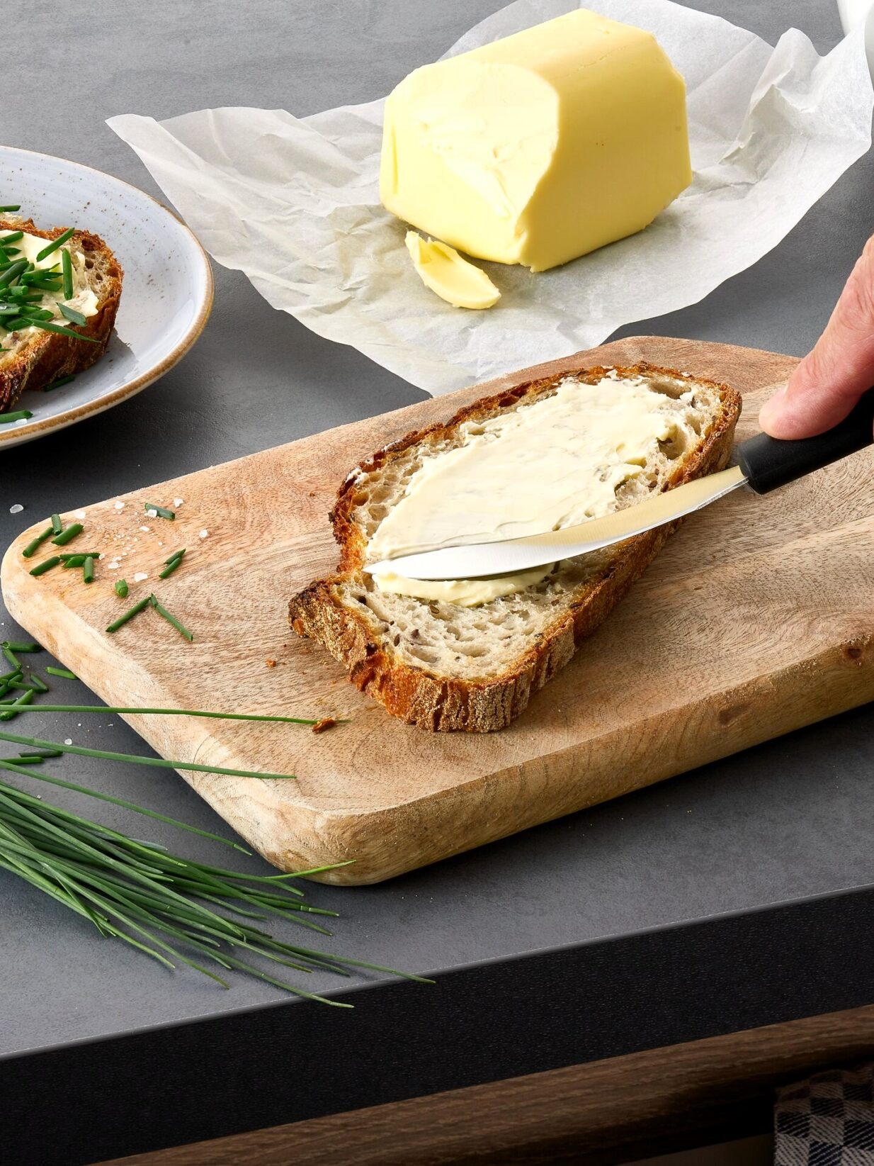 Tisch mit Holzbrettchen, auf dem eine Scheibe Brot liegt, die gerade mit Butter mithilfe des triangle Aufstrichmessers aufgetragen wird. Neben dem Brettchen befindet sich frischer Schnittlauch. Im Hintergrund ein weiteres belegtes Brot und ein Stück Butter.